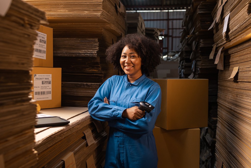 A woman holding an RF scanner standing among stacks of flat boxes and constructed boxes. 