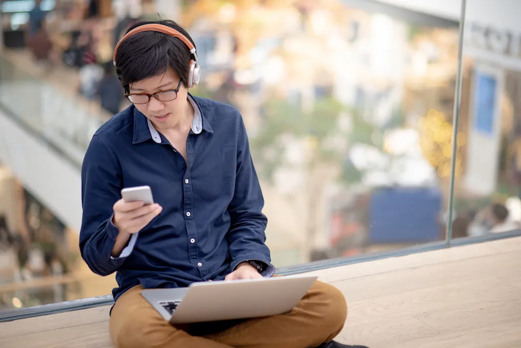 A young Asian male checking his phone while working at a computer.